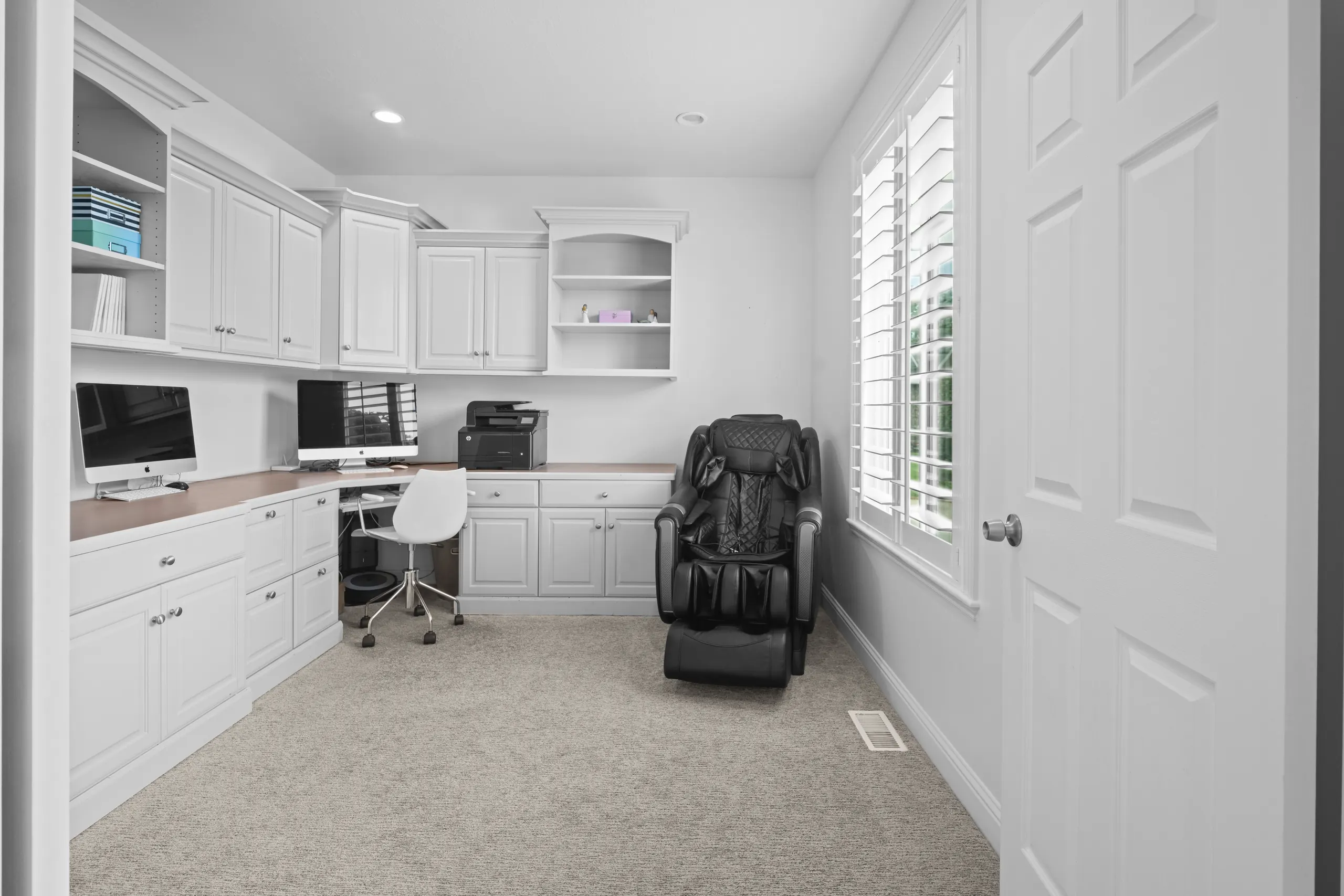 Home office lined with white built-in cabinetry and a corner desk, lit by a shuttered window