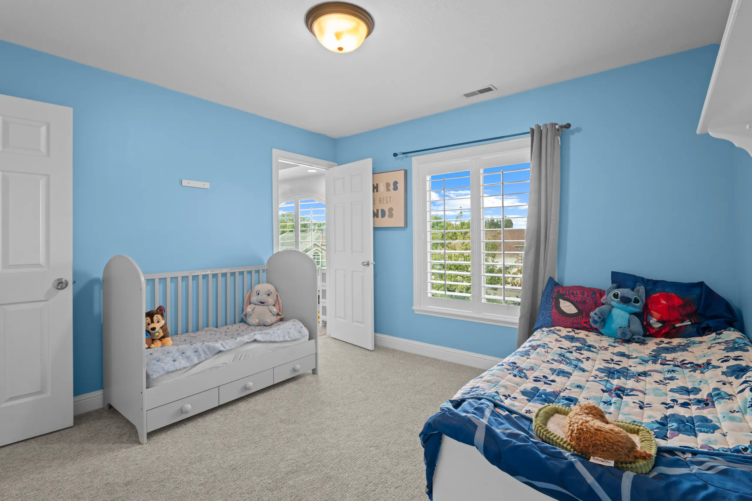 Children's bedroom painted sky blue with a toddler bed, a twin bed and a plantation-shuttered window